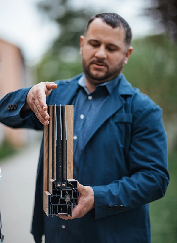 Man in a blue jacket holds a multi-chamber window frame cross-section outdoors.