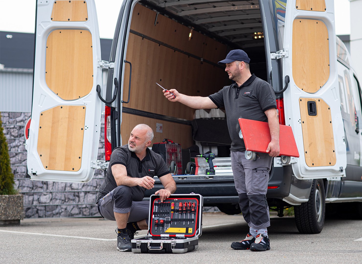 Two technicians work on a white cargo van with rear doors open; one kneels with an open tool case, the other holds a red board near the back.