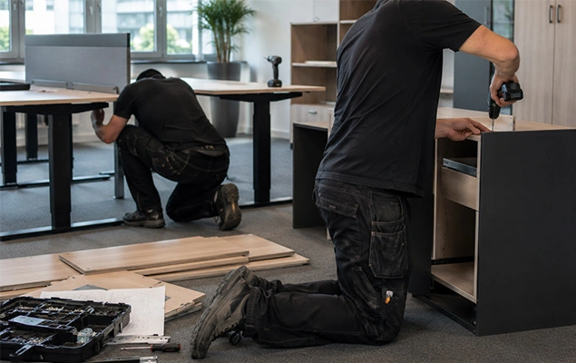 Two workers assemble dark wood office furniture; one kneels under a desk, the other drills a cabinet.