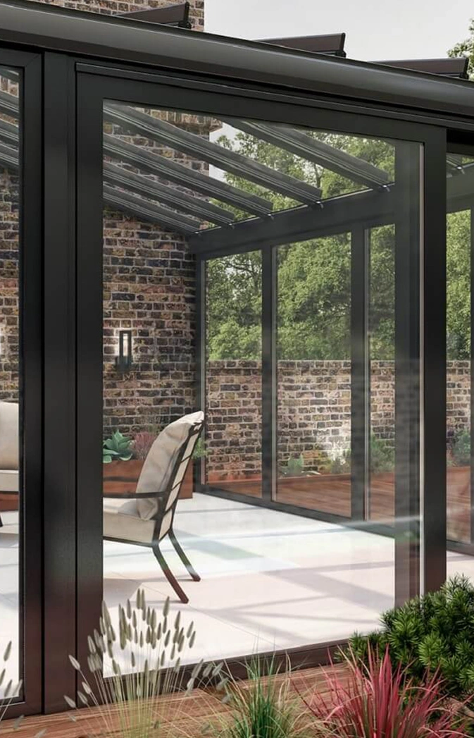 Modern glass sunroom with a black metal frame, brick wall, and a beige chair inside.